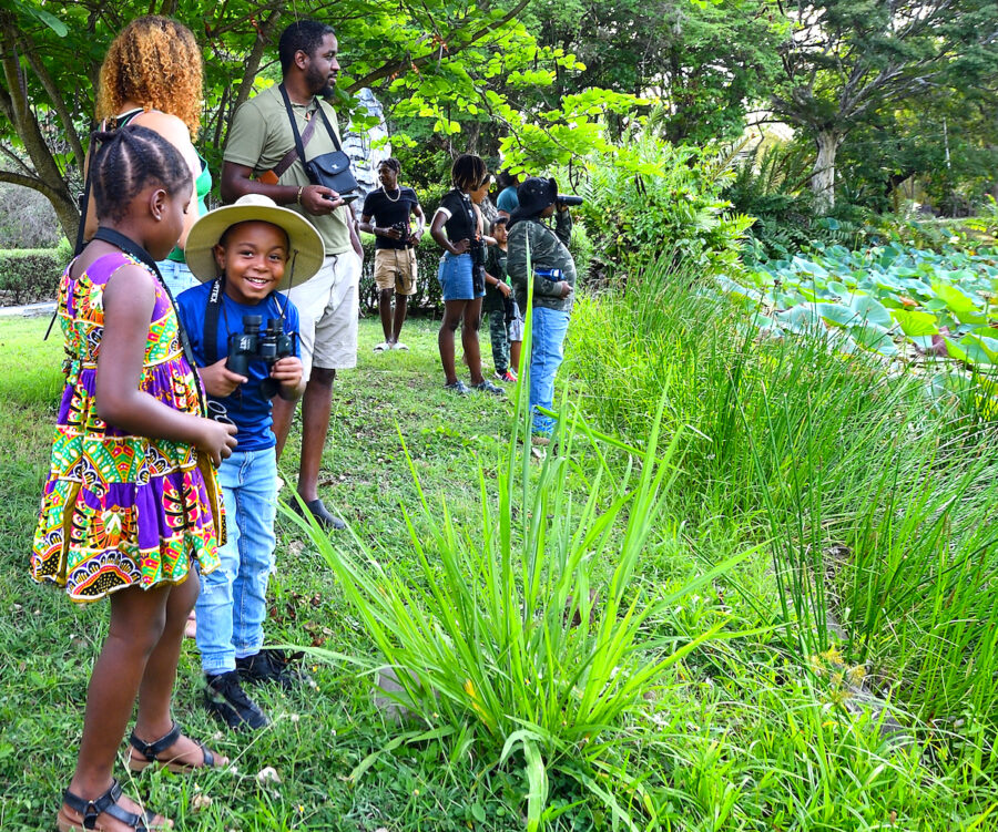 A group of adults and children birdwatching near a pond.