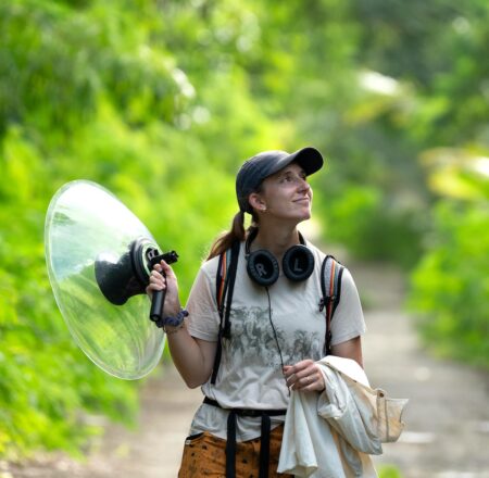Photo of woman on a forest trail holding sound recording device. 