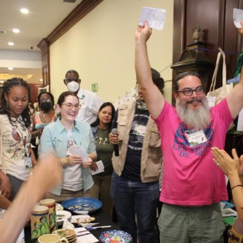 Group of people at an auction table. One man is holding his bid ticket in the air to indicate he has won the bid.